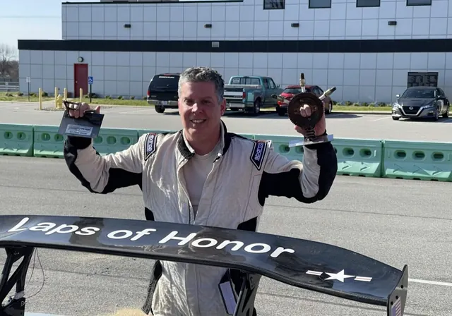 Driver in a race suit holding up two trophies behind a rear wing lettered Laps of Honor