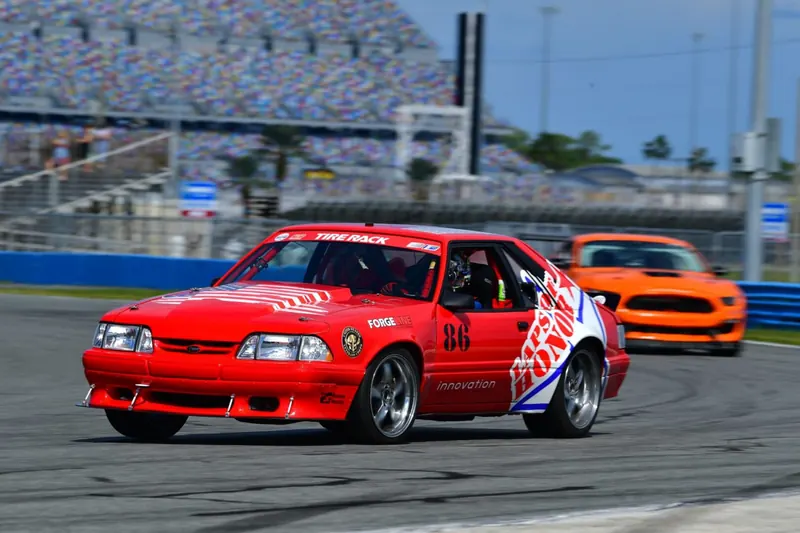 Red Fox-body Mustang in Laps of Honor livery racing on a speedway road course, an orange Mustang behind it
