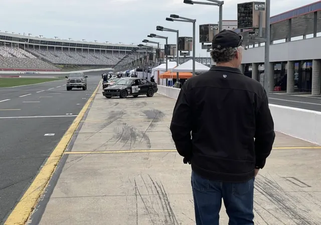 Team member standing in pit lane at a speedway, race cars on track beyond
