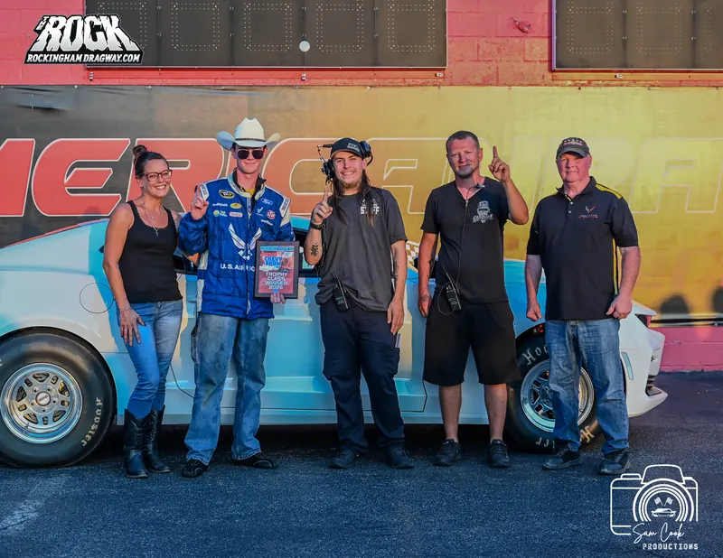 Hunter Marshall in a cowboy hat and blue racing jacket holding his Trophy Class Winner 2026 plaque, standing with four others in front of the white COPO Camaro at Rockingham Dragway