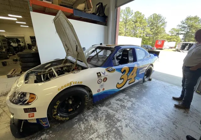 White and blue number 34 stock car with its hood up in the shop